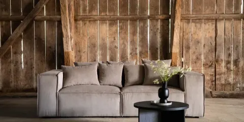 En stor, ljusbeige soffa med många kuddar står framför en rustik trävägg. Soffans mått är perfekta för ett rymligt vardagsrum.