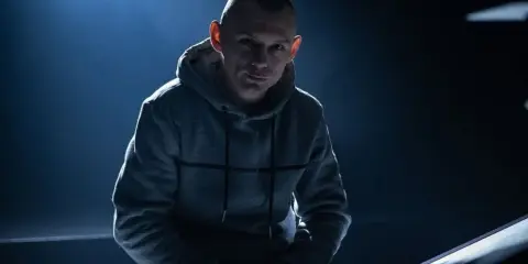Młody mężczyzna w szarej bluzie z kapturem, z ogoloną głową, w mrocznym świetle. Jego spojrzenie sugeruje opowieść o polskim rapie.