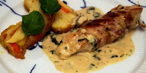 En rullad kyckling insvept i bacon, serverad med en krämig sås till kyckling och gratinerade potatisbitar med gröna blad.