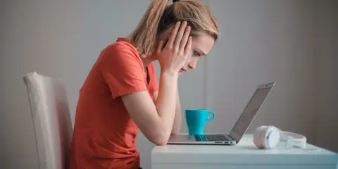 Een meisje met een korte spanningsboog staart naar haar laptop, handen aan haar hoofd. Een blauwe mok en koptelefoon staan naast haar.