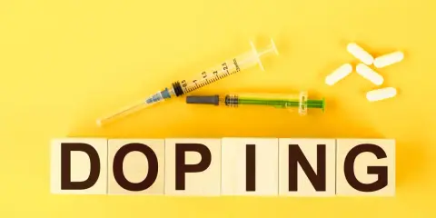 Dwa strzykawki i białe kapsułki na żółtym tle. Słowo "DOPING" napisane na drewnianych klockach.