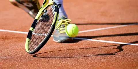 Piłka do tenisa ziemnego w locie, rakieta i buty gracza na ceglanym korcie.