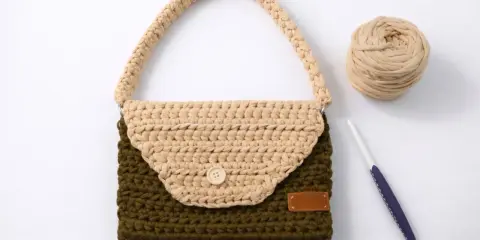 Bolso de trapillo bicolor, verde y beige, con asa y botón de madera. A su lado, ovillo de lana y ganchillo.