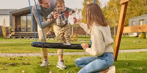 Radosne zabawy w ogrodzie: tata podtrzymuje dziecko na huśtawce, mama zachęca do zabawy.
