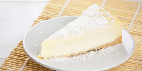 Une part de tarte noix de coco, crémeuse et généreusement saupoudrée de noix de coco râpée, repose sur une assiette blanche, prête à être dégustée.