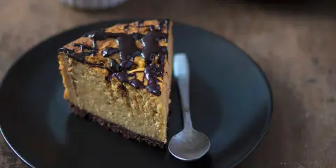 Tranche de cheesecake sans gluten sans lactose, nappée de chocolat noir, sur une assiette noire avec une cuillère.