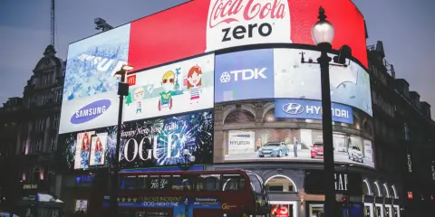 Wielkie ekrany reklamowe na placu, prezentujące różne rodzaje reklamy: Coca-Cola Zero, Samsung, Vogue, Hyundai, TDK. Widać też autobus i ludzi.