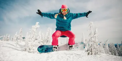 Dziewczyna w różowej czapce i goglach skacze na snowboardzie na tle zaśnieżonych drzew. To jest właśnie snowboard!