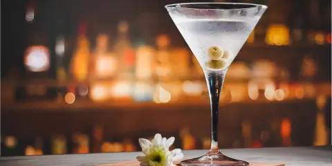 Martini extra dry z oliwkami, idealny do wieczornego relaksu. Na blacie stoi kieliszek z drinkiem i biały kwiatek.