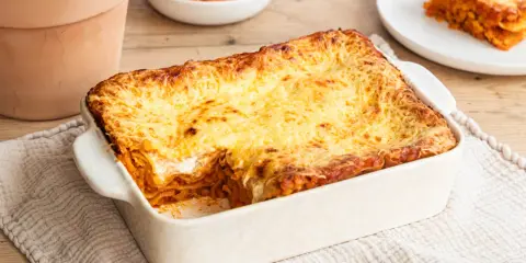 Herzhafte Linsen Lasagne mit goldbraun überbackener Käseschicht, daneben eine Schüssel roter Linsen und ein Teller mit einem Stück Lasagne.