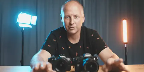 Mężczyzna z aparatami przed sobą, omawia podstawy fotografii. W tle niebieskie i pomarańczowe światła.
