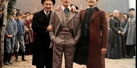 Trzech mężczyzn w eleganckich strojach, otoczonych tłumem, jakby w drodze do ziemi obiecanej.