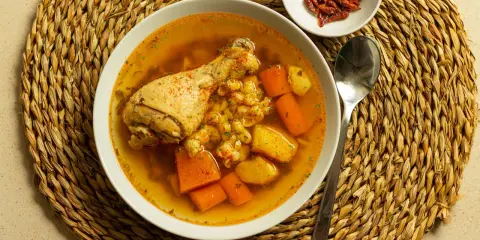 Un confortante brodo di pollo con pezzi di pollo, carote e patate, servito su una tovaglietta di paglia.