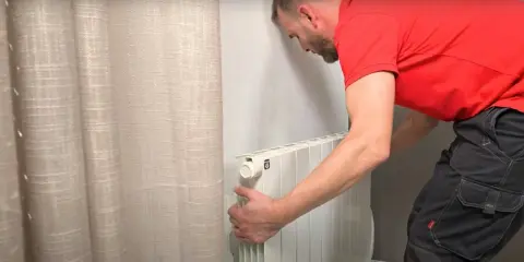 Un homme en t-shirt rouge et pantalon de travail ajuste un radiateur électrique blanc, prêt pour son branchement.