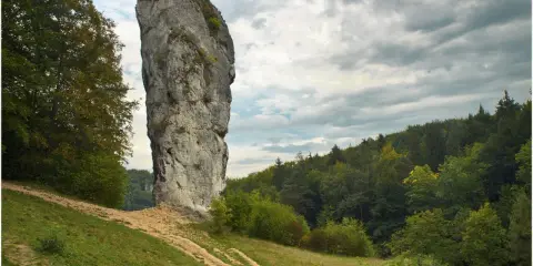 Imponująca skała, Maczuga Herkulesa, wznosi się na tle burzowego nieba. U jej podnóża zielona łąka i ścieżka prowadząca przez las.
