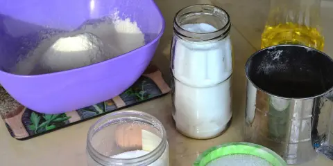 Filiżanka mąki, słoiki z cukrem, sitko i olej na blacie kuchennym.
