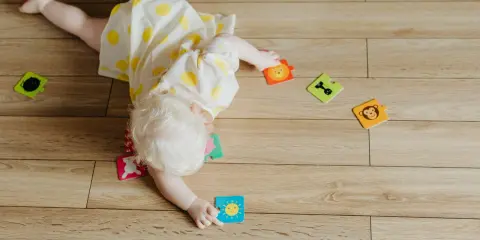 Dziecko w żółtej sukience z kółkami próbuje wykonać obrót z brzucha na plecy, otoczone kolorowymi puzzlami ze zwierzątkami.