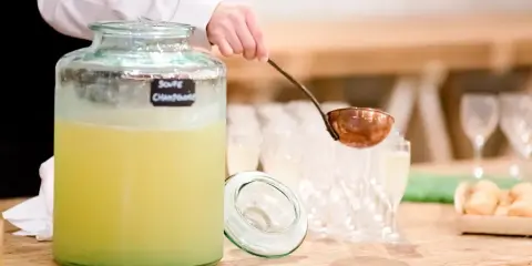 Une louche sert une soupe champenoise sans alcool dans un grand pichet en verre. Des verres à pied et des amuse-bouches sont disposés sur la table.