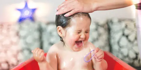 Ein Baby lacht und schreit vor Freude, während seine Haare gewaschen werden. Wasser spritzt, und es hält ein lila Band.