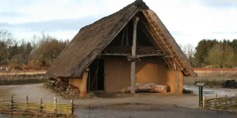 Ein rustikales Bauernhaus aus dem Mittelalter mit Strohdach und Lehmwänden, umgeben von Holzzäunen und Bäumen.