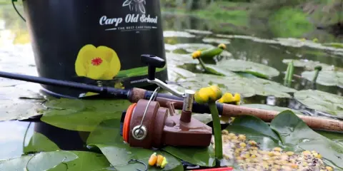 Wiadro "Carp Old School", wędka, kołowrotek i przynęta na liściach lilii wodnych. Idealne miejsce, by dowiedzieć się, jak łowić duże ryby na spławik.