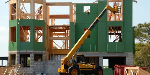 Chantier d'une maison individuelle en cours de construction. Une grue télescopique jaune est positionnée près de la structure en bois et des panneaux verts.
