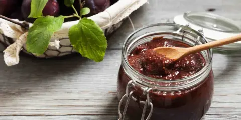 Słoik pełen domowych powideł śliwkowych z czekoladą, obok koszyk z dojrzałymi śliwkami.