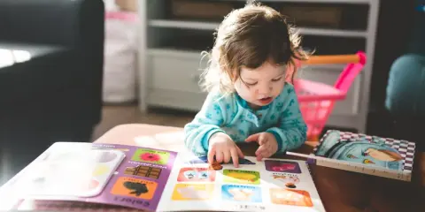 Mała dziewczynka z niepełnosprawnością intelektualną z zaangażowaniem przegląda książeczkę z obrazkami, wskazując paluszkiem na kolorowe ilustracje.