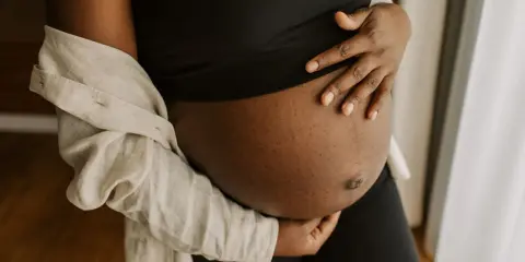 En gravid mage i tredje trimestern, varsamt omfamnad av händer. En ljus skjorta draperad över axeln.