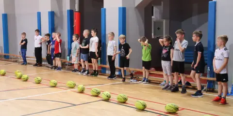 Dzieci w sali gimnastycznej czekają na rozpoczęcie zabaw ruchowych. Przed nimi leżą żółte piłki.