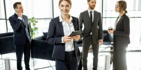 Une femme souriante, possiblement le chief of staff, tient une tablette. Autour d'elle, des collègues en costume discutent dans un bureau moderne.