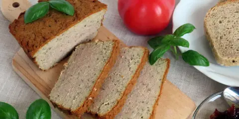 Pokrojony pasztet mieszany drobiowo-wieprzowy na desce, obok pomidor, chleb i konfitura.