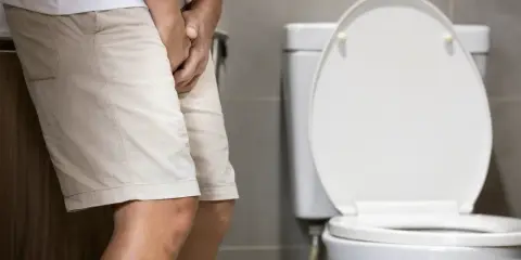 Mężczyzna w szortach stoi przy toalecie, trzymając się za krocze. Może to sugerować zatrzymanie moczu, które w ICD-10 ma określony kod.