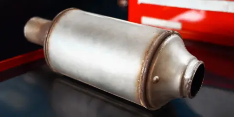 Metalowy tłumik, element układu wydechowego, leży na ciemnym tle. W tle widać fragment czerwonej skrzynki narzędziowej.