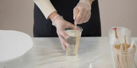 Hände mit Handschuhen mischen zwei komponenten harz in einem Becher. Kleine Farbtropfflaschen stehen bereit.