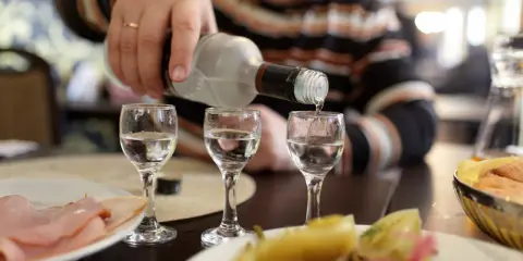 Ręka nalewa wódkę do kieliszków, przygotowując się na rekord promili. Na stole wędlina i przekąski.