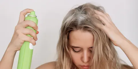 Frau sprüht sich Haarspray auf die Kopfhaut, was die Haare austrocknen kann.
