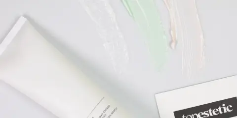 Biała tubka z kosmetykiem i kolorowe smugi kremów. Obok wizytówka z logo "topestetic", sugerująca znane marki kosmetyków.