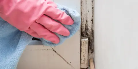 Mano con guanto rosa pulisce con panno azzurro la muffa rossa sul muro vicino a una cerniera.