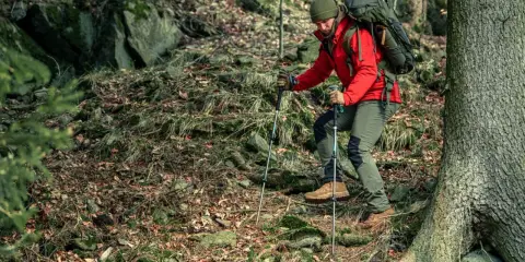 Mężczyzna w czerwonej kurtce i zielonej czapce wspina się po leśnym zboczu, używając kijków trekkingowych. To właśnie **outdoor** – życie na świeżym powietrzu.