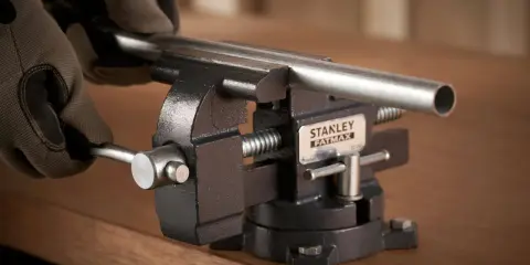 Manos enguantadas ajustan un tubo metálico en un tornillo de banco Stanley Fatmax, herramienta esencial para fijar piezas.