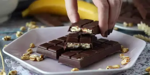 A hand breaks a chocolate bar filled with puffed rice and nuts on a pink plate.