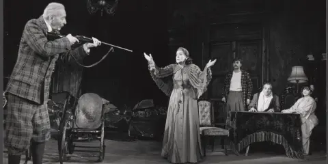 Scena z dramatu Mrożka. Starszy mężczyzna w kratę celuje z dubeltówki do kobiety z rozłożonymi rękami. Na scenie inni aktorzy.
