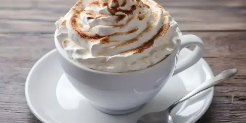 Ein cremiger Kapuziner Kaffee, üppig garniert mit Schlagsahne und Zimt, wartet auf einem Holztisch.