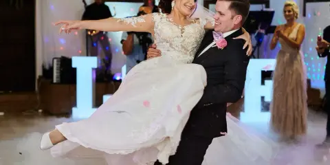 Pan młody niesie pannę młodą, tańcząc przy świetlnych literach LOVE. Idealny moment, by zastanowić się, co zaśpiewać na weselu dla pary młodej.