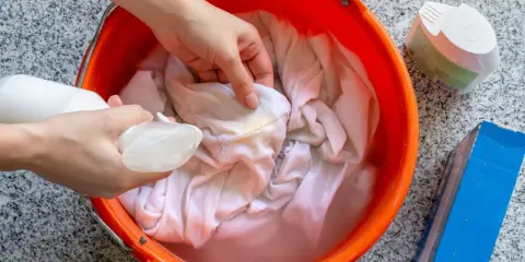 Ręce rozpylają płyn na poplamioną tkaninę w pomarańczowym wiadrze z wodą.