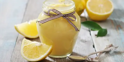 Tarro de lemon curd casero, decorado con un lazo, rodeado de limones frescos y rodajas.