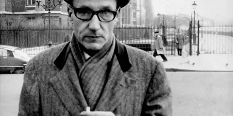 William Burroughs, con cappello e occhiali, tiene una sigaretta in mano, passeggiando in una strada urbana con persone sullo sfondo.