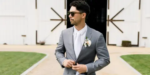 Man in a grey suit, white shirt, and tie, looking cool in sunglasses. Perfect for casual men's wedding guest attire in summer.