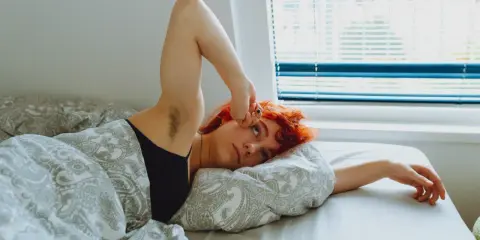 Jeune femme aux cheveux roux allongée dans son lit, le bras levé, le regard perdu. Elle semble fatiguée, peut-être un des symptômes manque magnésium.